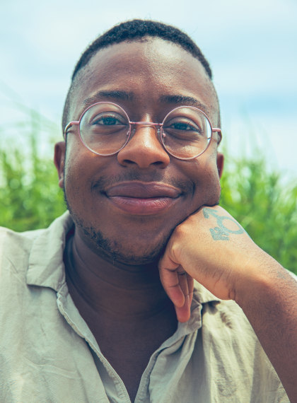 
	A Black man with cropped hair, shaved sides, and pink circular wire frames looks directly into the camera. They wear a cream linen button down and half smile. He's leaning his cheek on a closed fist with a trans symbol tattoo. The sky is blue and grass sprouts up behind him.