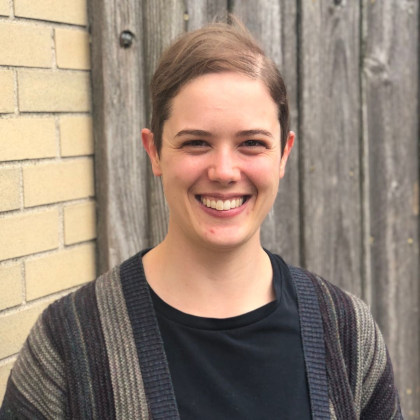 
		Alexis smiles into the camera. Alexis is a white person with short, fine, light brown hair parted to the side and brown eyes. Alexis wears a multi-colored knit sweater with subdued gold, purple, green and black vertical stripes, draped over a black shirt. In the background, a wooden gate with grey, vertical planks, adjoins a yellow brick wall. 
		