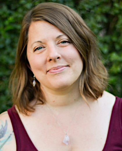 
		Annie Wiegand headshot: a middle aged white woman with shoulder-length brown hair and blue eyes. Annie is wearing a maroon-colored tank top and a purple amethyst necklace, with small beaded earrings. There is a hint of warm light coming from the right, and the background is blurred green foliage. 
		
		