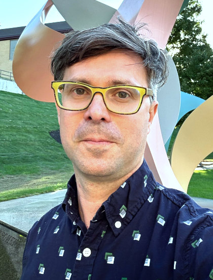 
		Colin Clark headshot. 
		Colin is white man with messy, graying brown hair and brown eyes. He is wearing highlighter-yellow glasses and a blue collared shirt. He’s standing outside in front of an elaborate sculpture composed of curving pink, yellow, blue, and orange metal spiraling above his head. 
		