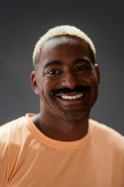 
Jerron Herman headshot. Jerron is a dark-skinned Black man with a big smile, dark moustache and bright blonde hair; he wears a soft yellow t-shirt. Photo Robbie Sweeny.
		