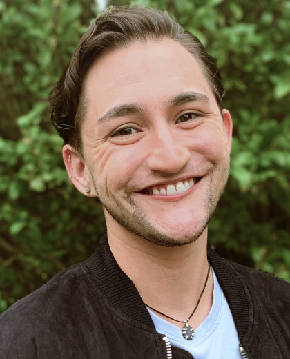 
		Kasson Marroquin headshot. 
		Kasson is a white person with swooped-back brown hair, 
		crinkled brown eyes, and a broad smile. He wears a casual t and jacket,
		and is in front of verdant green bushes.

		