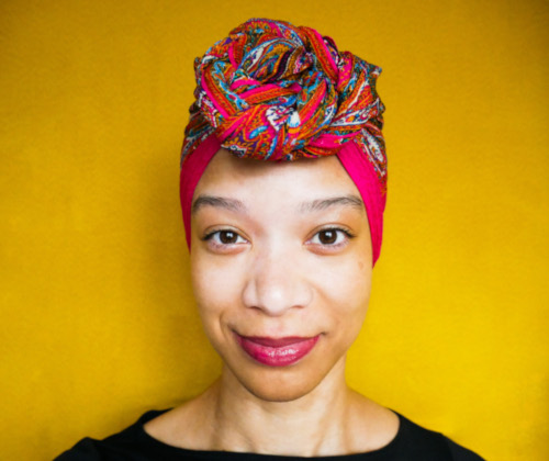 
Against a warm yellow background Morgan, a light-skinned Black and Indigenous woman looks directly at the camera with a slight knowing smile and sparkling brown eyes. A silk magenta headscarf with flashes of teal and purple is wrapped around her head with a large twisted knot in the front.
	