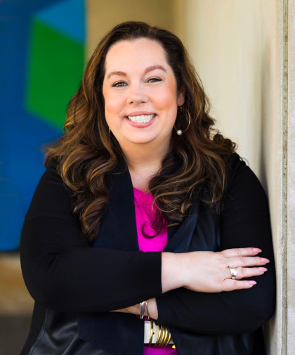 
		Mariclare Hulbert headshot. 
		Mariclare is a white woman with long brown hair, styled with curls. She smiles directly at the camera. She wears a bright pink top and a black jacket. Photo by Natalie Sinisgalli 
		