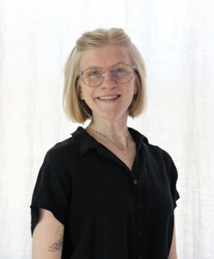 
 rachel smiles directly into the camera; she is a pale white woman with cropped light blonde hair and cornflower blue eyes. She wears acrylic-rimmed glasses and a black short sleeve button-up. Gold jewelry glints from her neck, nose, and dangles from her ears; a thin-lined tattoo of a wing peeks out from under her sleeve. photo credit: rachel hickman
	